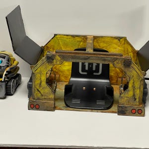May include: A yellow and black toy robot with the word "WALL-E" printed on it. The robot is standing next to a yellow and black toy truck with a black interior and a black seat. The truck has two red lights on the back. The truck has a black sign with the letters "11" on it.