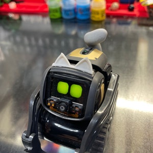 May include: A black and gold robot toy with a cat-like design. The robot has two large green eyes, a black camera lens, and white cat ears. The robot is on a black and gray base with wheels.