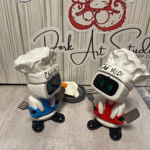 May include: Two robot chef figurines, each wearing a white chef's hat and apron, one blue and one red. They hold a spatula and knife, with a small pan of fried eggs between them. The text "Chef ALBI" is on the hat.