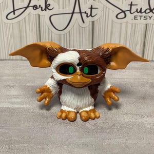 May include: A figurine of a Gremlin character, featuring brown and white fur, large ears, and green eyes. The figure has a mischievous expression and is posed with its arms outstretched. The figurine is a collectible item.