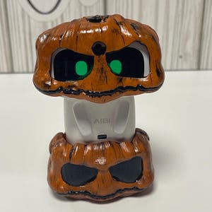 May include: A Halloween-themed decorative item featuring a pumpkin design. The orange pumpkin has black accents and green eyes. The base is white with the word "AIBI" printed on it. The item is designed to look like a jack-o'-lantern.