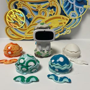 May include: A set of colorful plastic eggs with wings, each with a different pattern. The eggs are open and reveal a white interior. A white plastic figure with a black visor is in the center of the image.
