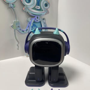 May include: A black robot toy with blue headphones, purple feet, and light blue and purple horns. The robot has a black screen and is standing on a white surface.