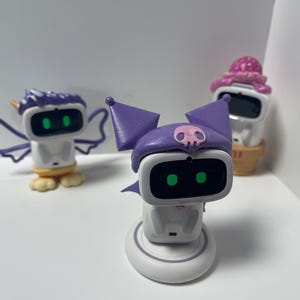 May include: Three small white robot toys with different accessories. The first robot has purple wings and a white unicorn horn. The second robot has a purple hat with a skull and crossbones design. The third robot has a pink and white ice cream cone hat.