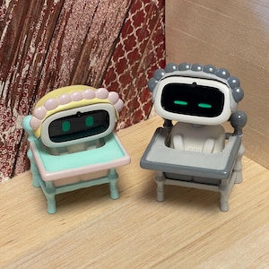 May include: Two toy robots, one in a pastel yellow and pink chair, the other in a gray chair. The robots have black screens with green horizontal lines for eyes. The chairs have rounded legs and are on a light wood surface.