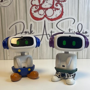 May include: Two robot figurines with white bodies, black screens, and blue and purple headbands. One robot has blue shorts and orange feet, the other has green shorts and black and white sneakers. The text "AIBI" is on the front of each robot.
