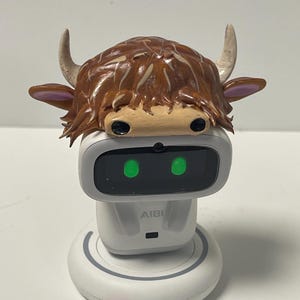 May include: A white AI robot with a cow-shaped headpiece. The cow head is brown with tan accents, horns, and ears. The robot has a black screen with two green lights and the text "AIBI" on the front.