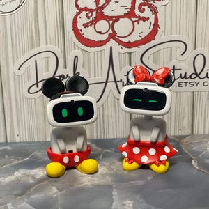 May include: Two white robot figurines, one with black ears and red shorts, the other with a red bow and a red skirt with white polka dots. The robots have black eyes and yellow feet. The background is a light wood grain.