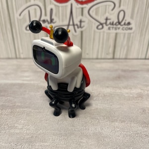 May include: A white and black robot figurine with a red cape and antenna-like features. The robot has a screen for a face and a small gold crown. The base is black with four legs. The text "Dork Art Studio ETSY.COM" is visible in the background.