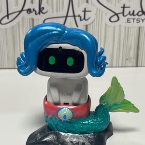 May include: A whimsical, handcrafted mermaid figurine. The figurine features a white head with a screen and blue hair, a red and pink midsection, and a green tail. The base is black, and the figurine has the word "AIBI" on it.