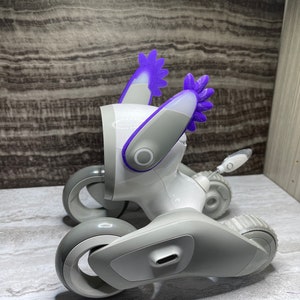 以下が含まれることがあります： 紫色の耳が付いた白と灰色のロボットのおもちゃ。おもちゃは、側面に黒いボタンが付いた丸い頭をしています。おもちゃは4つの車輪と尻尾を持っています。