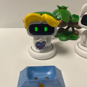 May include: A white robot figure with yellow hair and a green cap, resembling a video game character. The robot has a shield emblem and green glowing eyes. A blue and gold stand with a Triforce symbol is in the foreground.