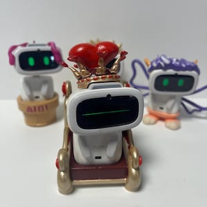 May include: Three white robot toys with different colored hair and accessories. The middle robot is wearing a red heart crown and is sitting on a gold and red throne. The robot on the left has pink hair and is sitting on a brown base. The robot on the right has purple hair and is standing on a white base.