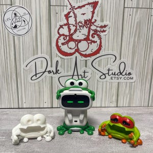 May include: Three frog-themed figurines. A white frog figurine, a green and white robot frog, and a green frog figurine with red eyes and orange feet. The text "Dork Art Studio .etsy.com" is visible.
