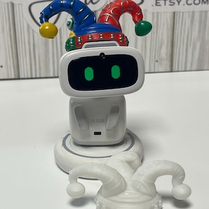 May include: A white AI robot toy with a colorful jester hat on top and a separate white jester hat. The robot has a screen with two green dots and the word "AIBI" on the front. The jester hats are blue, red, yellow, and green.