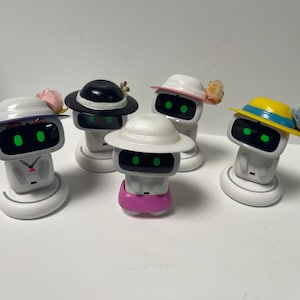 Op de afbeelding: Een verzameling witte robotfiguren, elk versierd met een hoed in een andere kleur en bloemaccenten. De hoeden zijn zwart, wit, geel en roze, wat een speels tintje geeft aan het ontwerp van de robots. De robots hebben groene ogen.