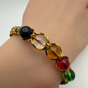 May include: A macrame bracelet featuring a series of colorful, round beads. The beads are black, clear, amber, red, and green, and are connected by a woven cord in shades of yellow, green, and brown. The text "FengShuiLuckyCharms" is visible.