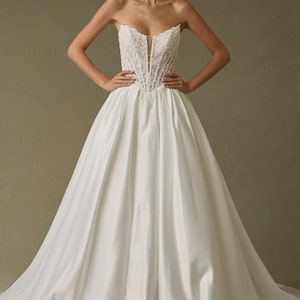 Peut inclure: Une robe de bal blanche sans bretelles dot&eacute;e d'un corsage structur&eacute; de style corset avec des d&eacute;tails en dentelle complexes et un d&eacute;collet&eacute; plongeant en V. La jupe est faite d'un tissu blanc lisse et uni, tombant en une silhouette ample et volumineuse.