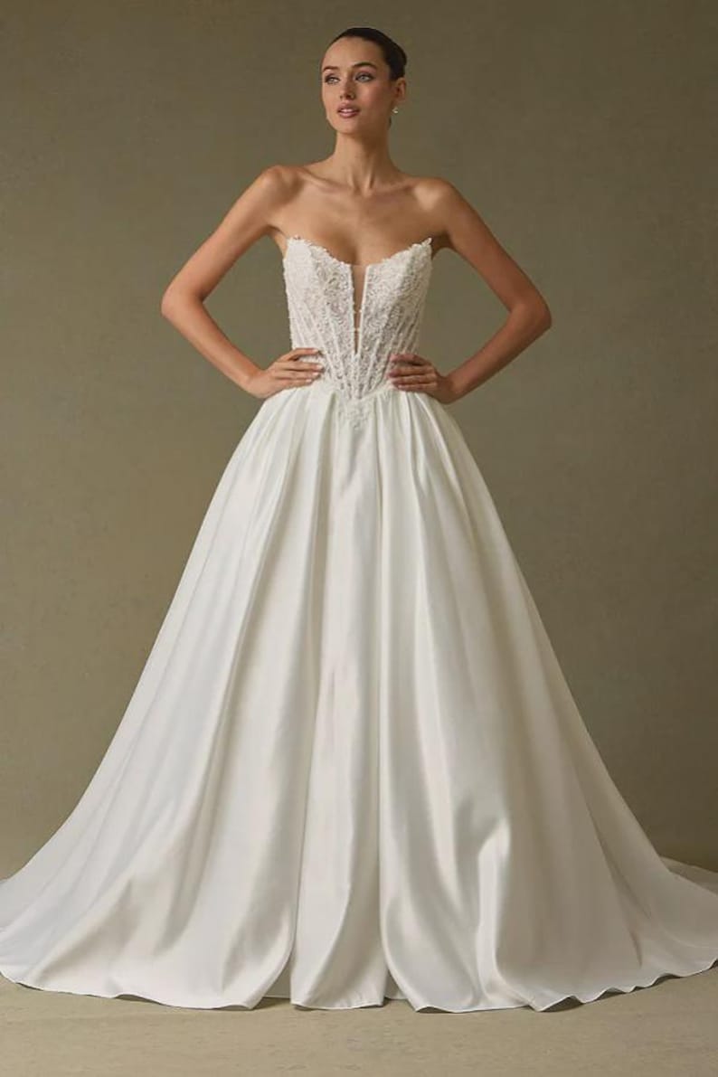 Peut inclure: Une robe de bal blanche sans bretelles dot&eacute;e d'un corsage structur&eacute; de style corset avec des d&eacute;tails en dentelle complexes et un d&eacute;collet&eacute; plongeant en V. La jupe est faite d'un tissu blanc lisse et uni, tombant en une silhouette ample et volumineuse.