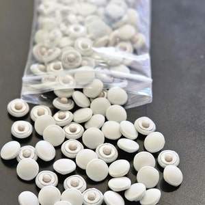 May include: A collection of white, round buttons with a fabric-covered top and a detailed center, spilling out of a clear plastic bag. The buttons are scattered on a dark surface, showcasing their uniform design and texture.