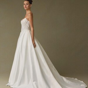 Peut inclure: Une robe de mari&eacute;e blanche longue jusqu'au sol avec un corsage bustier structur&eacute; orn&eacute; de d&eacute;tails en dentelle complexe et une jupe en satin lisse et ample qui se prolonge en une longue tra&icirc;ne. La robe pr&eacute;sente une taille ajust&eacute;e et une silhouette &eacute;pur&eacute;e.