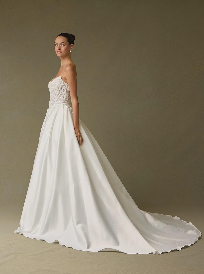 Peut inclure: Une robe de mari&eacute;e blanche longue jusqu'au sol avec un corsage bustier structur&eacute; orn&eacute; de d&eacute;tails en dentelle complexe et une jupe en satin lisse et ample qui se prolonge en une longue tra&icirc;ne. La robe pr&eacute;sente une taille ajust&eacute;e et une silhouette &eacute;pur&eacute;e.