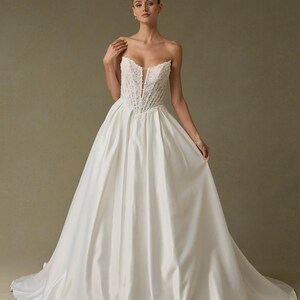 Peut inclure: Une robe blanche longue jusqu'au sol sans bretelles avec un corsage structur&eacute; style corset orn&eacute; de dentelle complexe et un d&eacute;collet&eacute; plongeant au centre. La jupe est en tissu blanc lisse et uni, s'&eacute;vasant en une silhouette volumineuse.