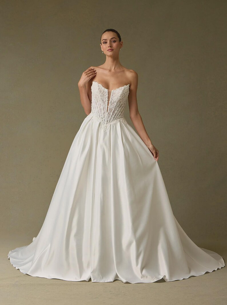 Peut inclure: Une robe blanche longue jusqu'au sol sans bretelles avec un corsage structur&eacute; style corset orn&eacute; de dentelle complexe et un d&eacute;collet&eacute; plongeant au centre. La jupe est en tissu blanc lisse et uni, s'&eacute;vasant en une silhouette volumineuse.