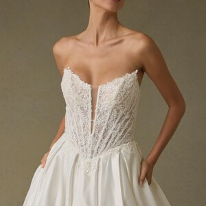 Peut inclure: Une robe de mari&eacute;e bustier ivoire dot&eacute;e d'un corsage style corset avec une broderie florale blanche complexe et un d&eacute;collet&eacute; plongeant avec un insert en maille transparente. La jupe est en satin lisse avec une silhouette ample et volumineuse.