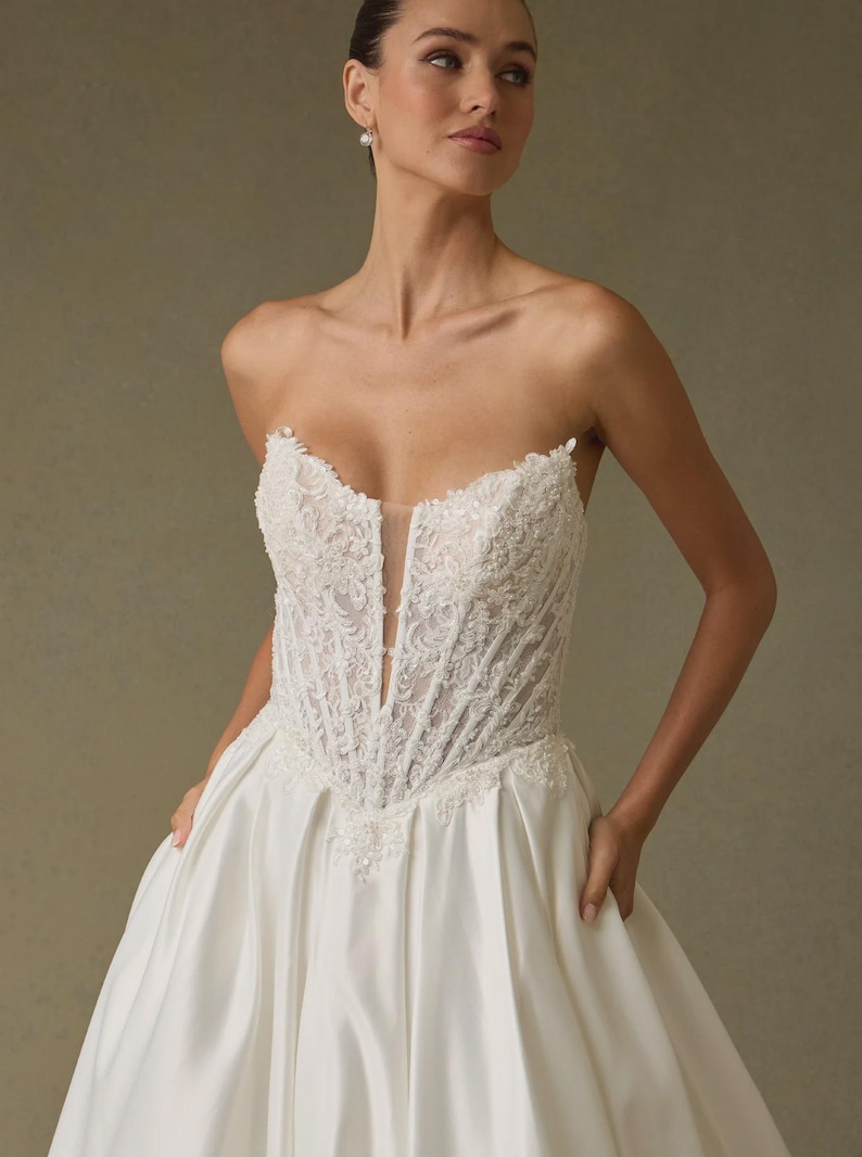 Peut inclure: Une robe de mari&eacute;e bustier ivoire dot&eacute;e d'un corsage style corset avec une broderie florale blanche complexe et un d&eacute;collet&eacute; plongeant avec un insert en maille transparente. La jupe est en satin lisse avec une silhouette ample et volumineuse.
