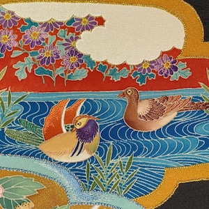 Op de afbeelding: Close-up van een decoratief textiel met twee mandarijneenden op een blauwe waterachtergrond. De eenden hebben een kleurrijk verenkleed, met oranje, gele en paarse accenten. De scène bevat rode en blauwe bloemelementen.