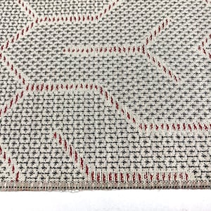 以下が含まれることがあります： 幾何学模様の織り生地のクローズアップ。生地はベージュで、黒いグリッド模様と赤い線が幾何学模様を形成している。