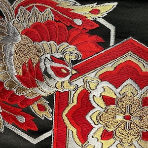 Pode incluir: Close-up de um tecido preto com bordados vermelhos, dourados e prateados. O design apresenta um pássaro estilizado com uma grande crista de penas e padrões florais intrincados.