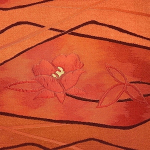 Könnte beinhalten: Ein leuchtend orangefarbener Stoff mit einem gewebten Muster aus dunkelbraunen, geschwungenen Linien und gestickten Blumenmotiven. Die zentrale Blume ist in Rottönen mit einem blassgelben Zentrum gehalten, ergänzt durch ein dreiblättriges Zweigdesign.