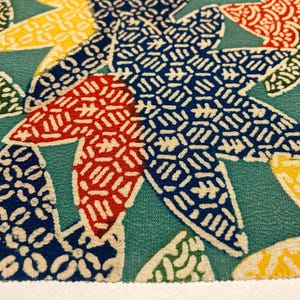 May include: Close-up of a fabric featuring a repeating pattern of stylised maple leaves. The leaves are in shades of red, yellow, blue, and green, set against a teal background. The design has a traditional, decorative aesthetic.