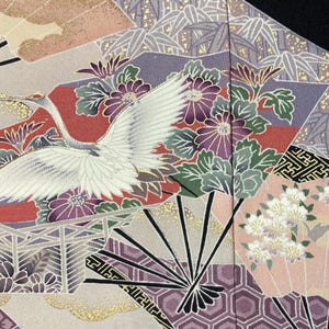 May include: Close-up of a patterned fabric featuring a white crane in flight, surrounded by floral and geometric designs. The fabric incorporates shades of purple, red, pink, and gold, with fan and bamboo motifs.