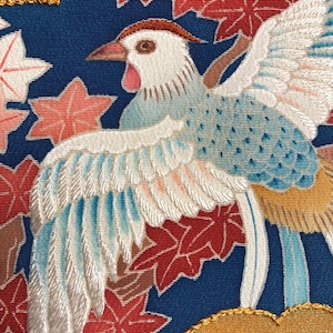 Könnte beinhalten: Nahaufnahme eines bestickten Textils mit einem Vogel mit weißen, blauen und roten Federn. Der Vogel ist vor einem dunkelblauen Hintergrund mit roten und rosa Ahornblättern platziert. Die Stickerei hat ein detailliertes, strukturiertes Aussehen.