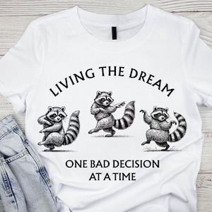 May include: White t-shirt with a black and white design of three raccoons in different poses. The text "LIVING THE DREAM" is above the raccoons, and "ONE BAD DECISION AT A TIME" is below. The shirt is made of white fabric.