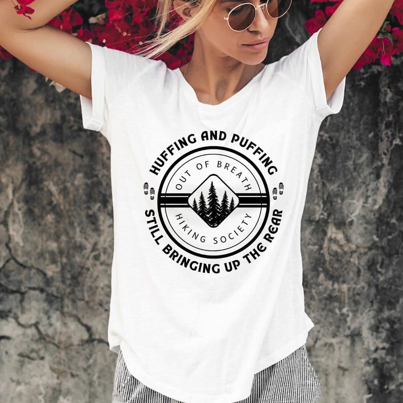 May include: White t-shirt with a circular black graphic that reads "Huffing and Puffing Out of Breath Hiking Society Still Bringing Up The Rear." The graphic features a mountain scene. The shirt has short sleeves.