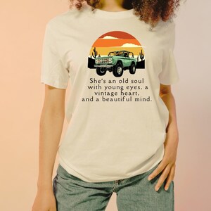 May include: Cream-colored t-shirt featuring a graphic of a vintage green SUV set against a sunset backdrop with cacti. The text reads, "She's an old soul with young eyes, a vintage heart, and a beautiful mind."