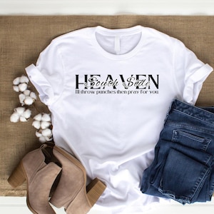 Może przedstawiać: Biała koszulka z czarnym nadrukiem: "Heaven South Side I'll throw punches then pray for you". Koszulka leży na brązowej powierzchni, obok pary brązowych botków i niebieskich dżinsów.
