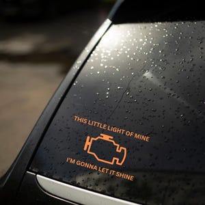 Może przedstawiać: Czarny samochód z naklejką na tylnej szybie. Naklejka przedstawia stylizowaną pomarańczową ikonę kontrolki silnika i tekst "This little light of mine, I'm gonna let it shine."