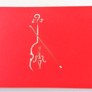 May include: A red paper card with a white cutout of a cello and bow. The cello is in the center of the card and the bow is angled across the cello.
