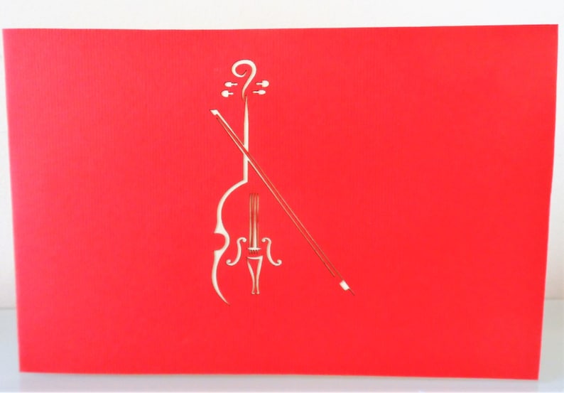 May include: A red paper card with a white cutout of a cello and bow. The cello is in the center of the card and the bow is angled across the cello.