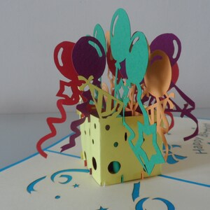 Puede incluir: Una tarjeta pop-up 3D con una caja amarilla llena de globos y serpentinas de colores. La tarjeta dice "Feliz cumplea&ntilde;os".