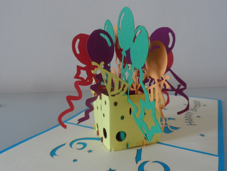 Puede incluir: Una tarjeta pop-up 3D con una caja amarilla llena de globos y serpentinas de colores. La tarjeta dice "Feliz cumplea&ntilde;os".