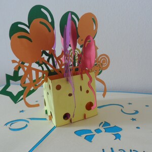Puede incluir: Una tarjeta pop-up amarilla y blanca con un dise&ntilde;o de bloque de queso. La tarjeta presenta un dise&ntilde;o 3D de globos y confeti. La tarjeta est&aacute; abierta y el interior es blanco con una cinta azul y estrellas.