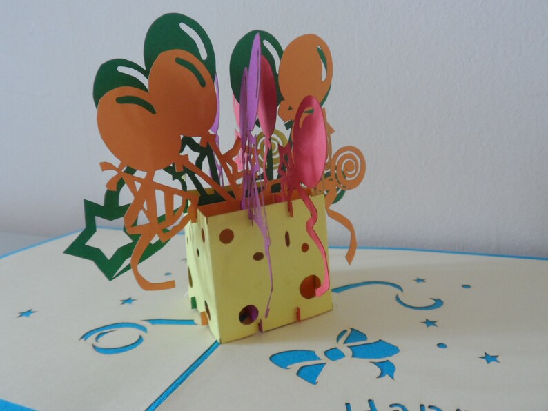 Puede incluir: Una tarjeta pop-up amarilla y blanca con un dise&ntilde;o de bloque de queso. La tarjeta presenta un dise&ntilde;o 3D de globos y confeti. La tarjeta est&aacute; abierta y el interior es blanco con una cinta azul y estrellas.