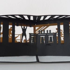 Peut inclure: Carte pop-up en papier noir et dor&eacute; repr&eacute;sentant une sc&egrave;ne de salle de sport. La carte pr&eacute;sente des silhouettes de personnes s'entra&icirc;nant avec des poids et d'autres &eacute;quipements de gym. La carte est ouverte, r&eacute;v&eacute;lant l'int&eacute;rieur.