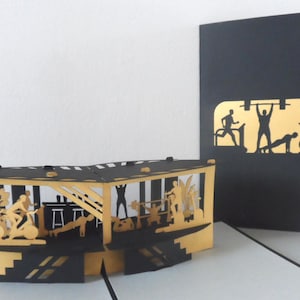 Pode incluir: Um cartão de felicitações pop-up com um tema de ginásio, feito de recortes de papel dourado e preto. O cartão mostra pessoas a exercitar-se com pesos, passadeiras e bicicletas de exercício. Ao lado, um cartão preto com uma silhueta dourada de ginásio.